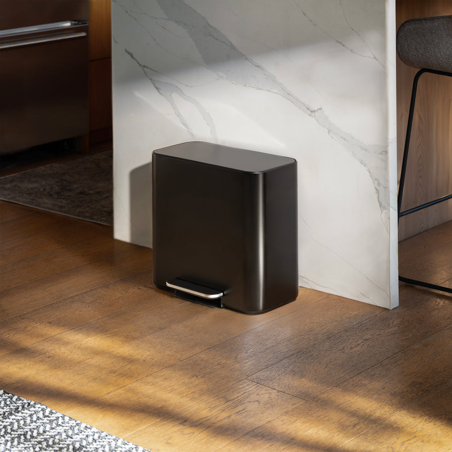 Simplehuman's 16L Matte Black Mini Step Can with pedal on wood flooring in a kitchen with a marble countertop behind it.