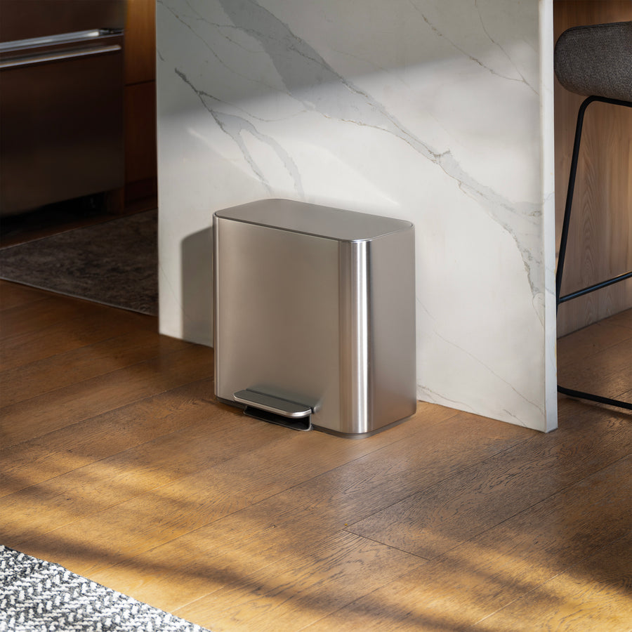 Simplehuman's 16L Brushed Stainless Steel Mini Step Can with pedal on wood flooring in a kitchen with a marble countertop behind it.