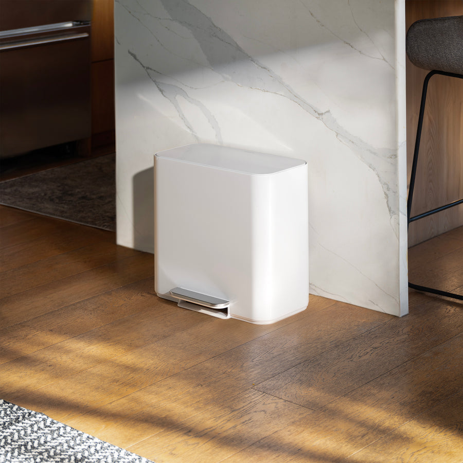 Simplehuman 16L White Mini Step Can with foot pedal, positioned on wood flooring next to a black storage cabinet with books next to it and plants on the other side.
