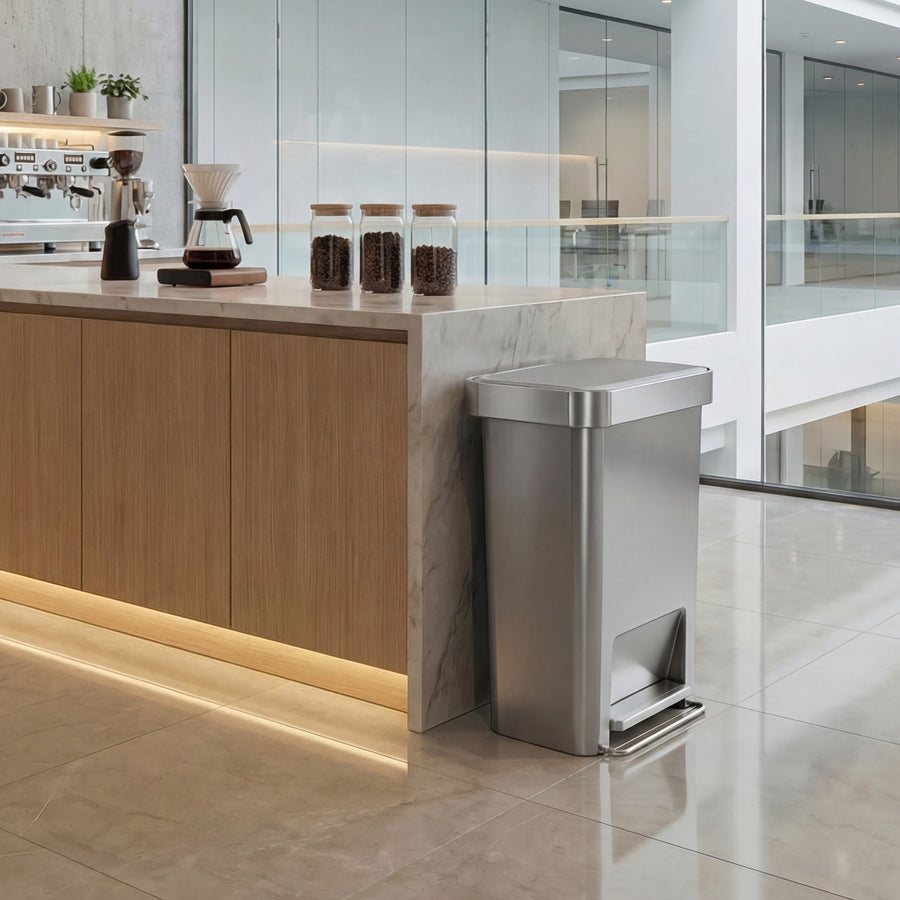 Simplehuman's 45L Hybrid Step Can in Alloy in an office coffee area setting. It is placed against a well-lit marble counter top with wooden drawers. 