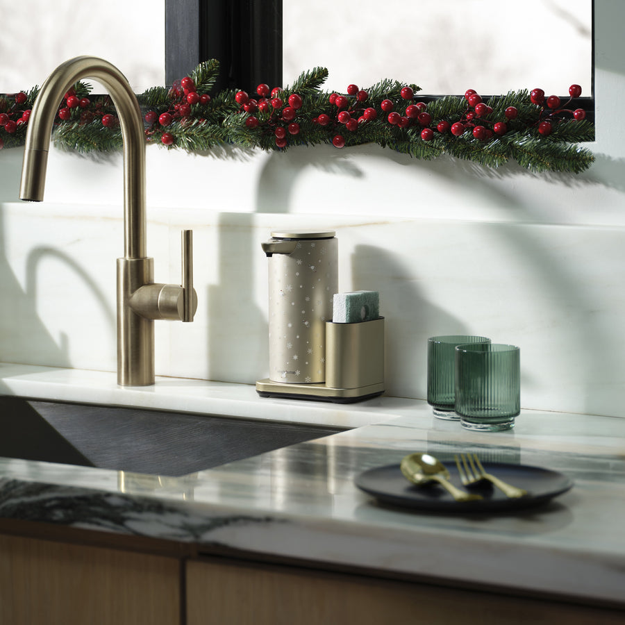 Modern kitchen sink with gold appliances, marble countertop, and a angled Simplehuman Matte Gold 14 oz. Liquid Sensor Pump, decorated with a Simplehuman 2025 Holiday Sleeve. Green glass cups and festive greenery with red berries line the window.