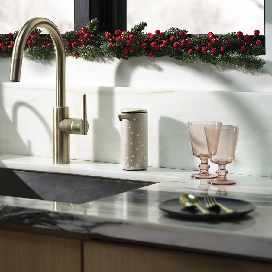 Modern kitchen sink with gold appliances, marble countertop, and an angled view of the Simplehuman Matte Gold 9 oz. Liquid Sensor Pump, decorated with a Simplehuman 2025 Holiday Sleeve. Green glass cups and festive greenery with red berries line the window.