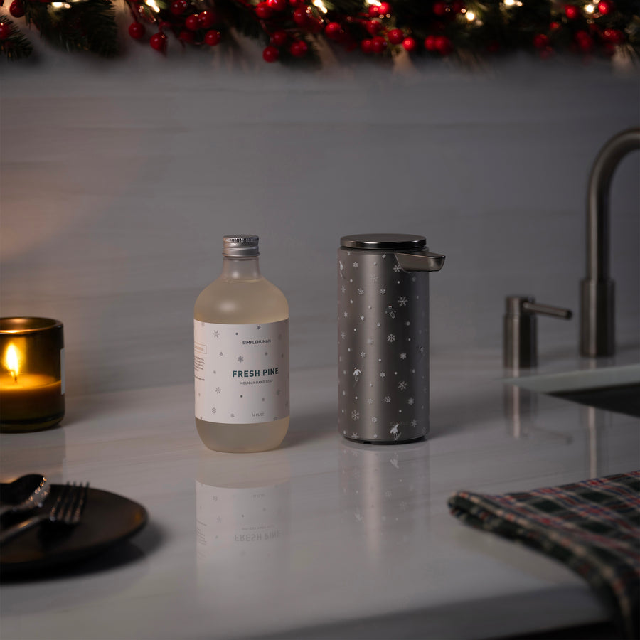 Holiday garments with warm mood lighting including Simplehuman's 9 oz. Liquid Sensor Pump in their 2025 Limited Edition Holiday Sleeve next to their new bottled Holiday Hand Soap in the scent Fresh Pine on a countertop.
