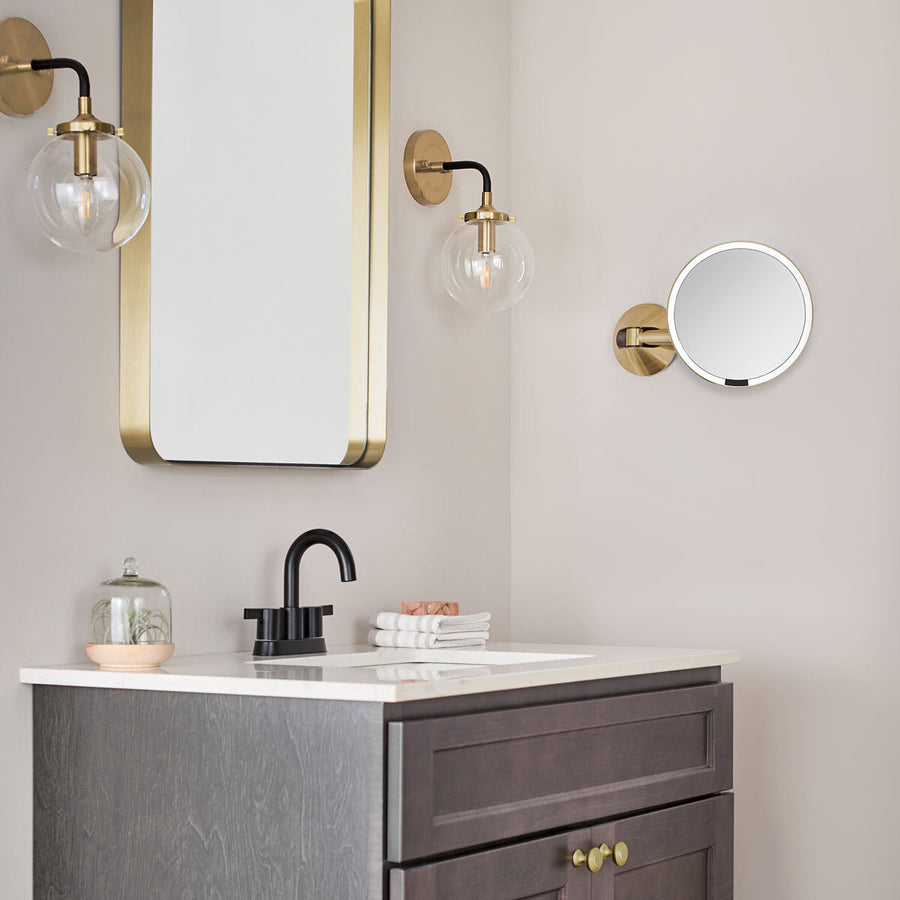Lifestyle view of Simplehuman Wall Mount Sensor Mirror in Brass finish over a dark wood vanity, touch-free light.