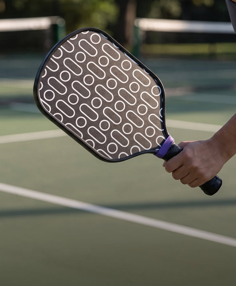 A close-up of a pickleball paddle being held by a person on an outdoor pickleball court. The paddle has a dark gray face with a white geometric pattern, a black edge, and a purple grip wrap.