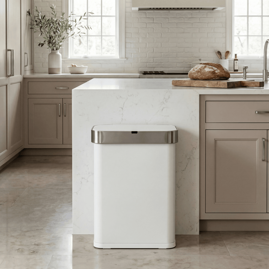 Simplehuman's 58L Sensor Can in White in a modern kitchen setting with neutral cabinetry, white marble counter tops and walls, with slight greenery and natural light in the background.